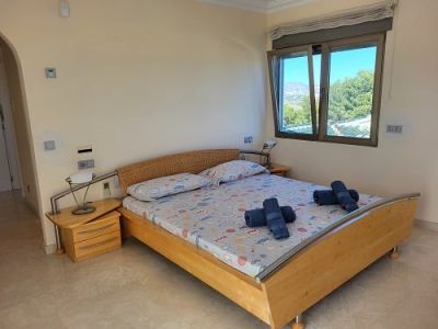 Dormitorio luminoso con cama de madera clara, cabecero de mimbre y mesita de noche. La ventana abierta ofrece vistas a la exuberante vegetación y las montañas.