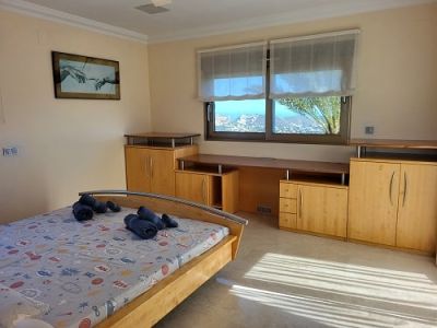 Dormitorio luminoso con cama de madera clara, escritorio integrado y armarios. Las ventanas ofrecen vistas panorámicas a la costa, iluminando la estancia con luz natural.