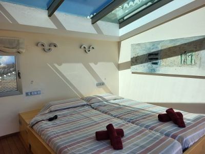 Dormitorio luminoso con dos camas individuales, ropa de cama a rayas y toallas rojas. Destaca un tragaluz que inunda la estancia de luz natural y un cuadro abstracto en la pared.