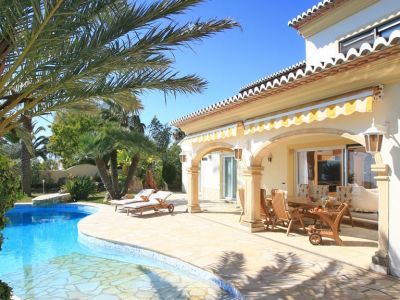 El exterior de la villa presenta una piscina de forma libre y una amplia terraza de piedra que conduce a una naya cubierta con mobiliario de comedor de madera y toldos rayados.