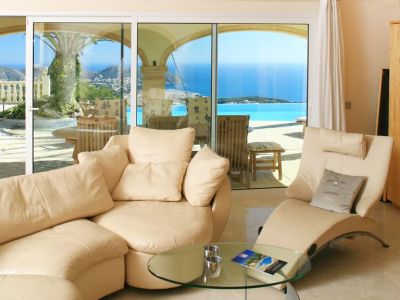 Espaciosa sala de estar con sofás curvos en tonos claros y suelo de mármol, ofreciendo acceso directo a la terraza con piscina infinity y vistas panorámicas al mar Mediterráneo.