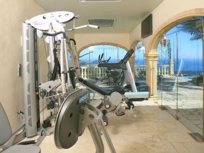 Gimnasio privado con equipo Hoist y Life Fitness, suelos de mármol y ventanales arqueados con vistas panorámicas al mar y la piscina. Claraboyas aportan luz natural.