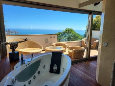 Jacuzzi interior con suelo de madera que se abre a una terraza privada amueblada con ratán, ofreciendo vistas panorámicas al mar Mediterráneo.