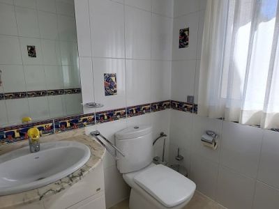 Luminoso baño con azulejos blancos, cenefa decorativa colorida y piezas de acento. Cuenta con un tocador de mármol con lavabo integrado, inodoro moderno y ventana con cortina traslúcida.