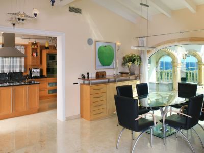 Luminoso comedor con mesa de cristal y sillas negras, suelos de mármol pulido. Grandes arcos acristalados ofrecen vistas panorámicas al mar y la montaña, conectando con una cocina moderna.