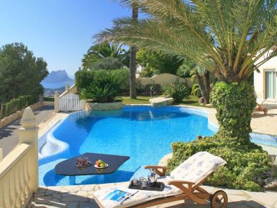 Piscina curva con una mesa negra sumergida y refrescos, junto a una terraza de piedra con tumbona. Se aprecian vistas lejanas al mar y una formación rocosa bajo un cielo despejado.