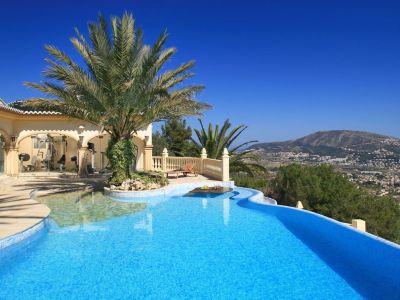 Piscina curva de mosaico azul con isla de palmeras y mesa sumergida. Gimnasio acristalado y terraza con vistas panorámicas a la montaña.