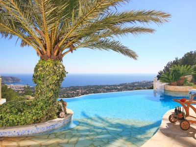 Piscina infinita con vistas panorámicas al mar y la costa, destacando una palmera cubierta de hiedra y un carrito de bar de madera en el solárium de piedra.