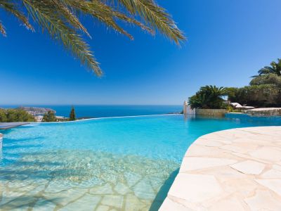 Piscina infinity con entrada tipo playa y bordes de piedra clara, ofreciendo vistas panorámicas al Mediterráneo y la costa, bajo un cielo azul.