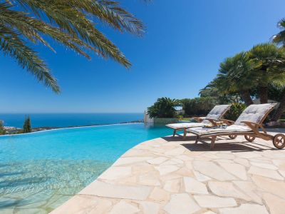 Terraza con piscina infinita y dos tumbonas de madera sobre un patio de losas, con amplias vistas al mar Mediterráneo y la costa lejana.