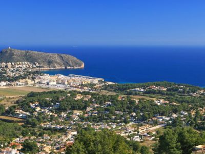 Vista panorámica desde una colina, mostrando un pueblo costero, un puerto deportivo, una montaña y el mar Mediterráneo bajo un cielo despejado.