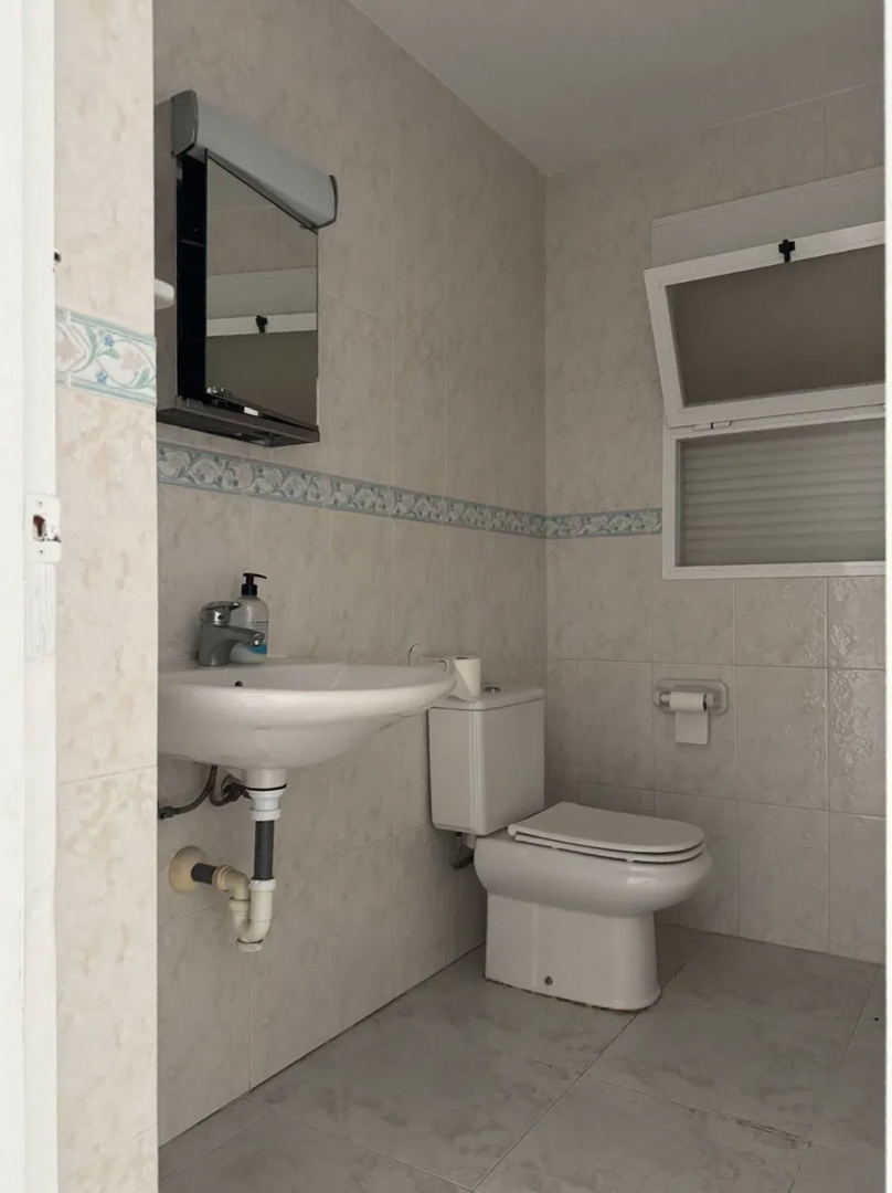 Baño funcional con azulejos claros y cenefa decorativa. Incluye lavabo de pared, inodoro y espejo con luz, complementado por una ventana abatible para ventilación.