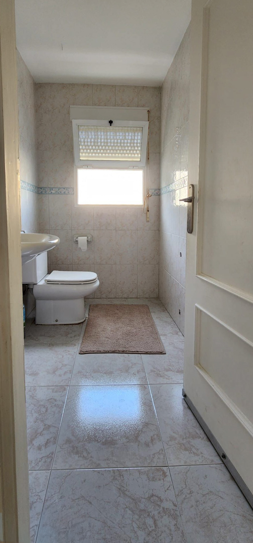 Baño funcional con inodoro, lavabo y ventana. Presenta azulejos de pared y suelo en tonos crema con un detalle decorativo azul, y una alfombrilla.