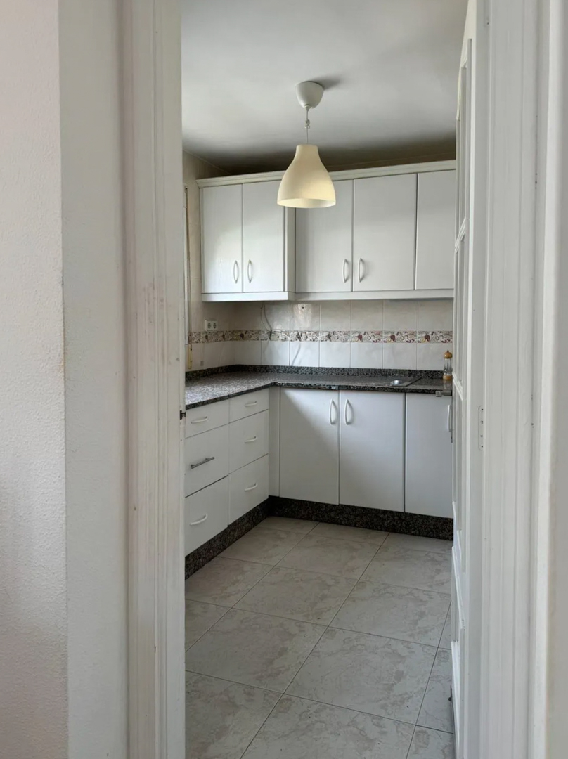 Cocina funcional con armarios blancos, encimera de granito oscuro y azulejos decorativos. Un colgante de techo ilumina el espacio, ofreciendo un lienzo para modernizar.