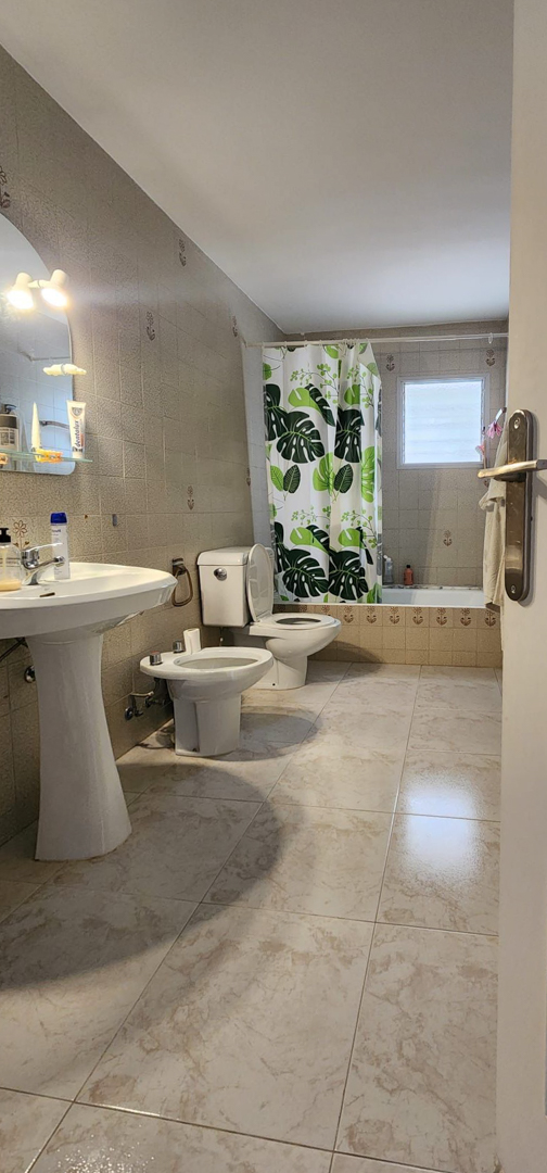 Cuarto de baño funcional con suelo de baldosas de efecto mármol claro y paredes alicatadas en tonos beige con detalles. Incluye lavabo de pedestal, inodoro, bidé y bañera con cortina estampada y ventana esmerilada.