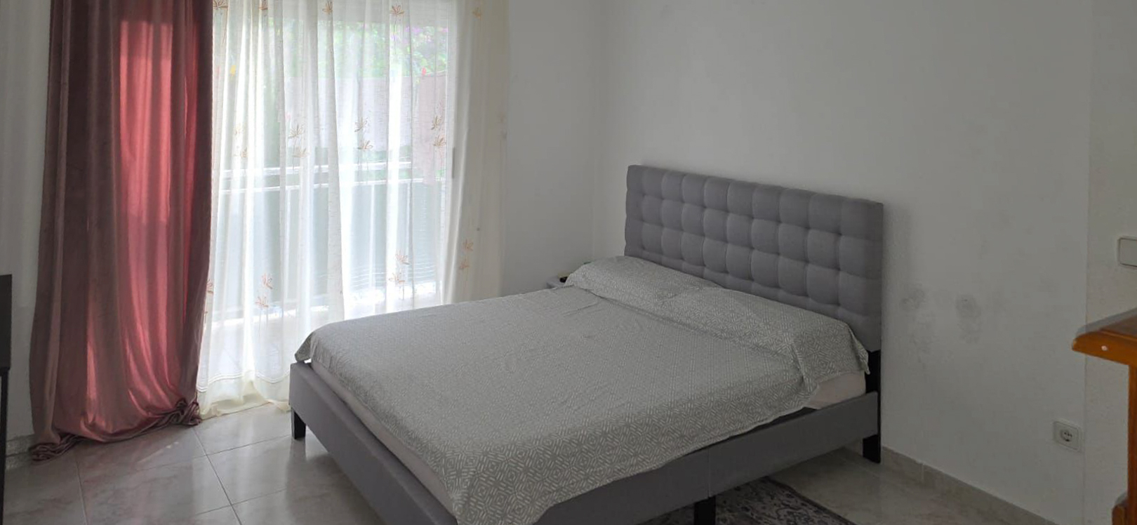 Dormitorio con cama tapizada gris y cabecero capitoné, con ropa de cama clara. Luz natural entra por el gran ventanal con visillos y cortinas granates, sobre suelo de baldosas.