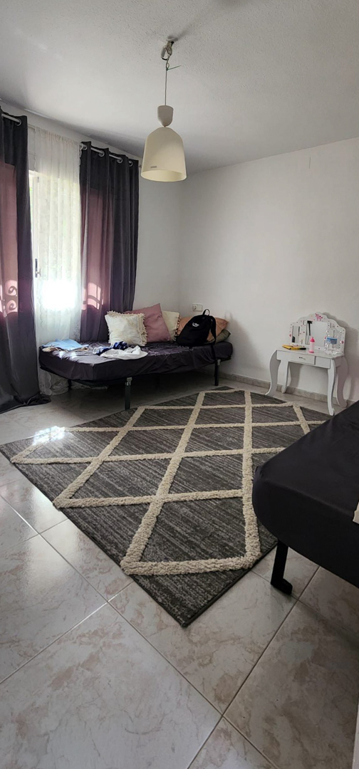 Dormitorio con suelo de baldosas claras, una alfombra geométrica gris y un diván. La ventana con cortinas oscuras permite la entrada de luz natural, junto a un tocador blanco.