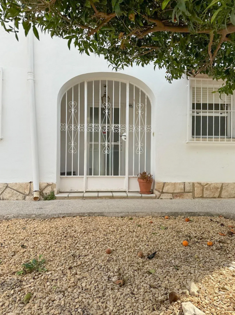 Entrada principal de la villa con un portón de metal arqueado y seguro. El jardín de grava frontal muestra frutos caídos y un árbol frutal que sombrea la fachada.