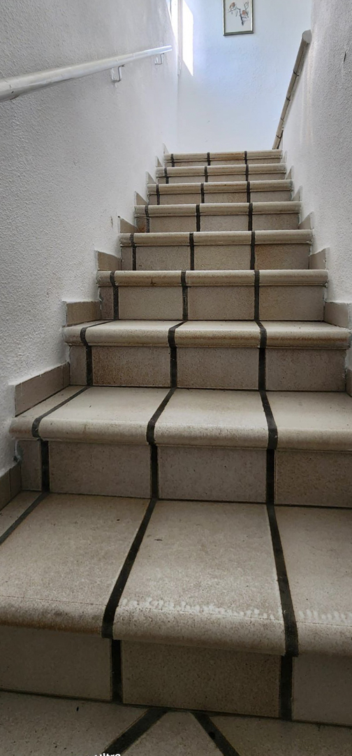Escalera interior con peldaños de azulejos claros y juntas oscuras, paredes blancas texturizadas y barandillas sencillas. Luz natural ilumina la parte superior.