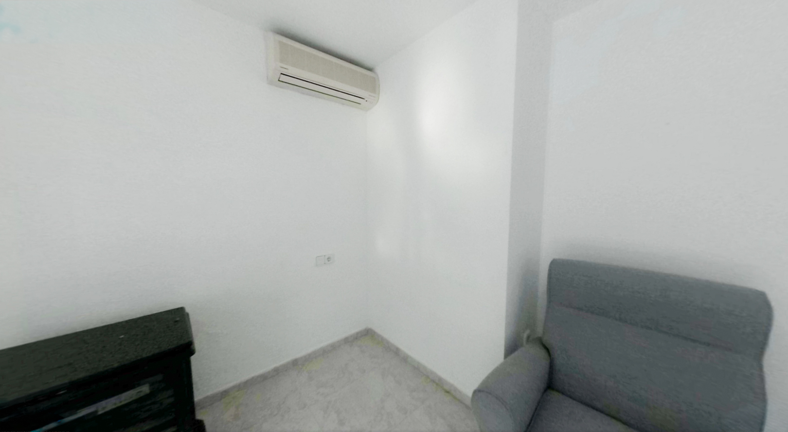 Esquina de una habitación con paredes blancas y suelo de baldosas claras. Dispone de aire acondicionado split y un enchufe en la pared, junto a un sillón gris y un mueble oscuro.