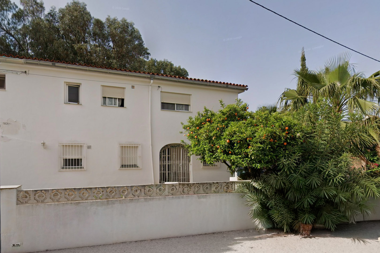 Fachada principal de la villa con paredes blancas, tejado de teja roja y ventanas con rejas. Destaca un naranjo cargado de fruta y exuberante vegetación en el jardín.