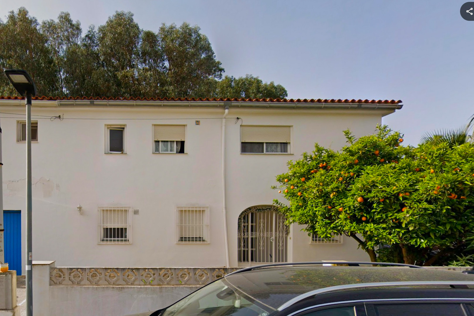 La fachada blanca de la villa de dos plantas muestra un tejado tradicional, una entrada arqueada con reja ornamental y ventanas con rejas. Un naranjo maduro, cargado de frutos, se alza prominentemente.
