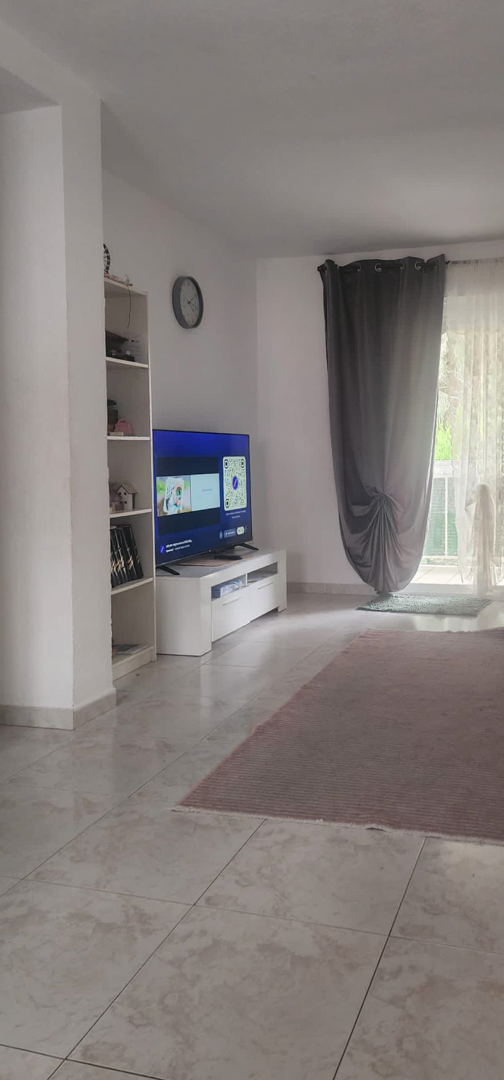 La sala de estar presenta suelo de baldosas claras y paredes blancas, con un moderno mueble de TV y estantería. Luz natural entra desde el acceso a la terraza, con cortinas grises y visillos blancos.