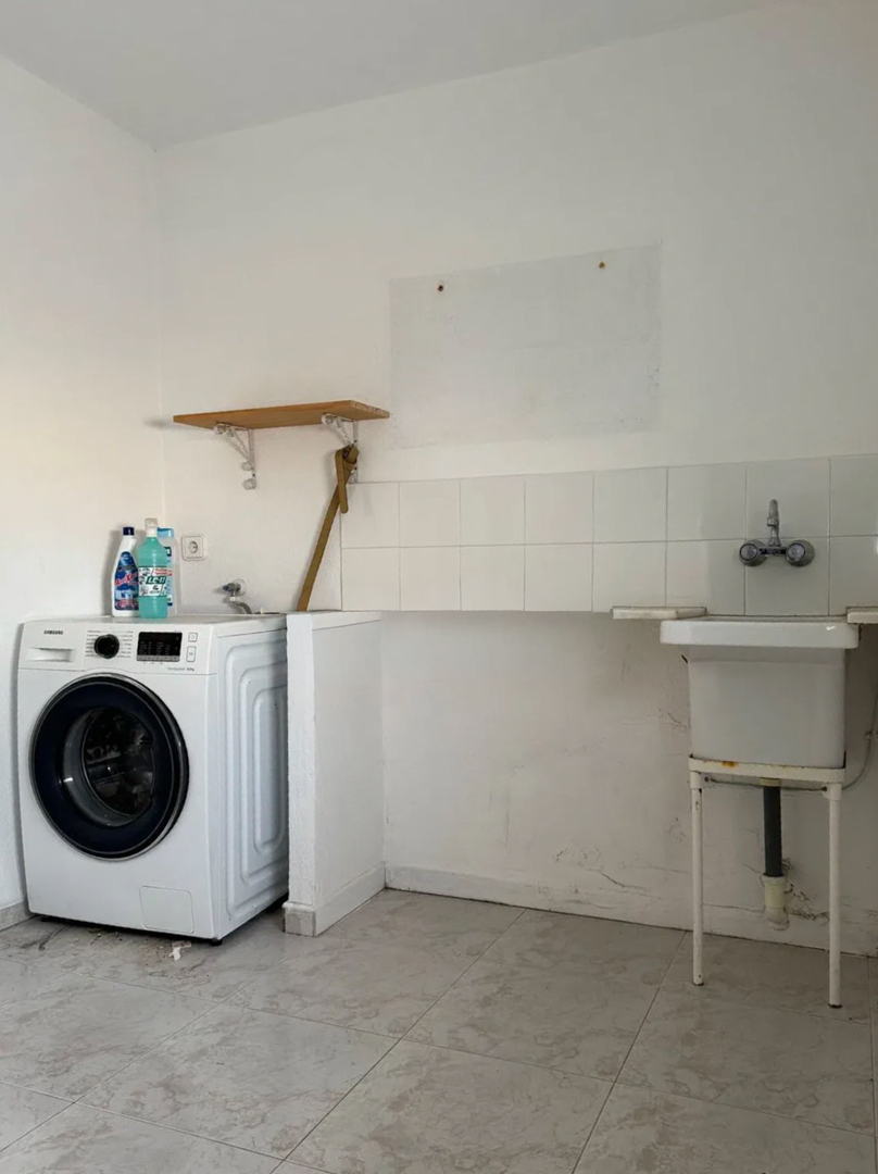 Lavadero funcional equipado con lavadora, fregadero de pila y azulejos blancos en la pared. El suelo es de baldosas claras y cuenta con una estantería.