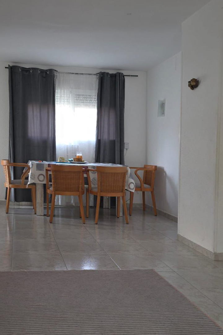 Luminoso comedor con mesa de madera y cuatro sillas de mimbre, junto a una ventana con cortinas grises. El espacio presenta suelo de baldosas claras y una alfombra rayada.