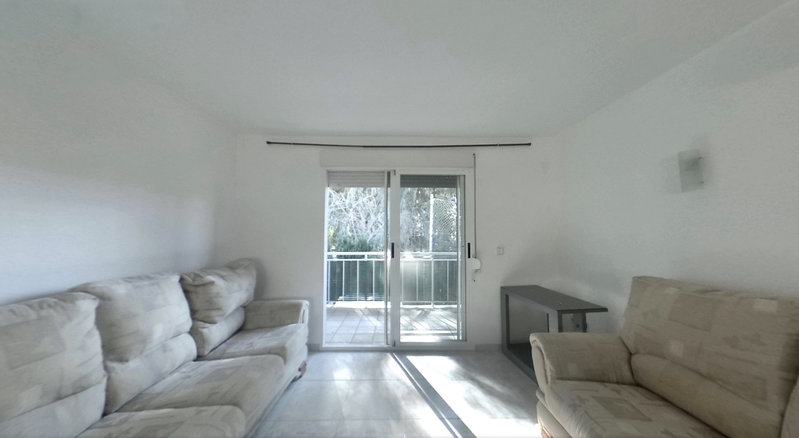 Luminoso salón con sofás claros, suelo de baldosas y puertas correderas de cristal que dan a un balcón, inundando el espacio con luz natural.