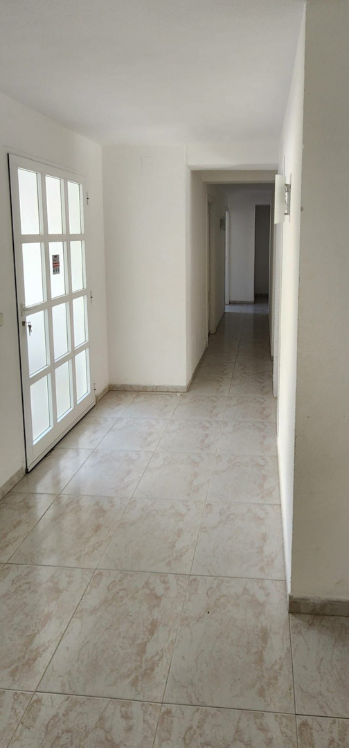 Pasillo interior con suelo de baldosas claras y paredes blancas. A la izquierda, una puerta con paneles de vidrio esmerilado; a la derecha, un aplique de pared ilumina el largo corredor.