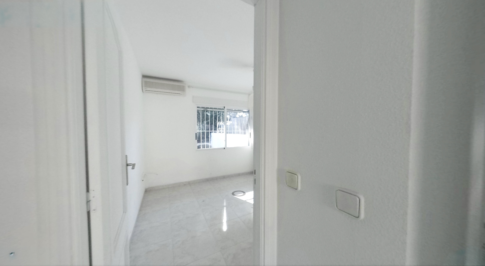 Vista de un dormitorio vacío con suelo de baldosas claras, paredes blancas y un aparato de aire acondicionado. La ventana con rejas de seguridad permite la entrada de luz natural.