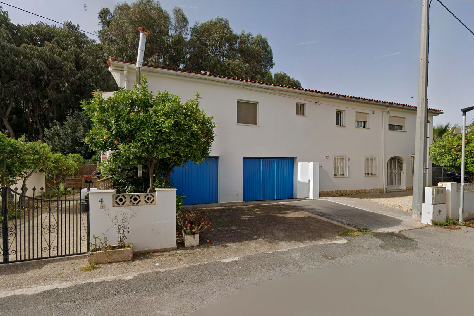 Vista frontal de la villa pareada con fachada blanca, dos puertas de garaje azules y un frondoso naranjo junto a la verja de hierro forjado que da acceso a la propiedad.