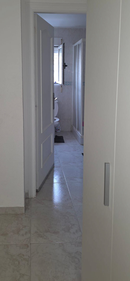 Vista parcial de un baño con suelo de gres claro, inodoro, ventana y ducha con mampara. Azulejos de pared con cenefa decorativa.