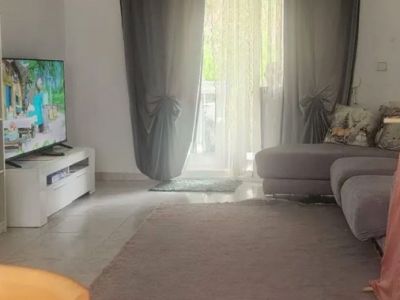 Amplio salón con suelo de baldosas claras, una alfombra neutra y un sofá modular gris. La luz natural entra por la puerta acristalada, con cortinas visillo y grises, que da acceso a una terraza exterior.