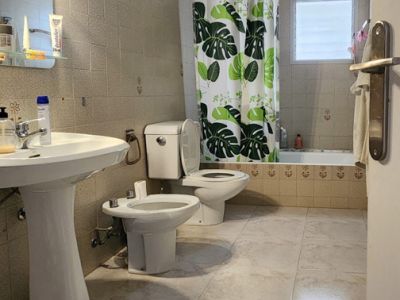 Baño con azulejos de tonos claros, lavabo de pedestal, inodoro, bidé y bañera con cortina de ducha de hojas tropicales y ventana esmerilada.