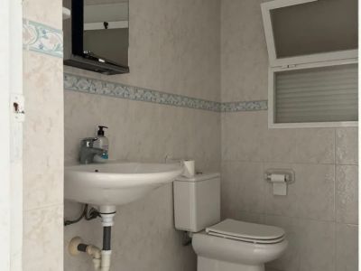 Baño funcional con azulejos claros y cenefa decorativa. Incluye lavabo de pared, inodoro y espejo con luz, complementado por una ventana abatible para ventilación.