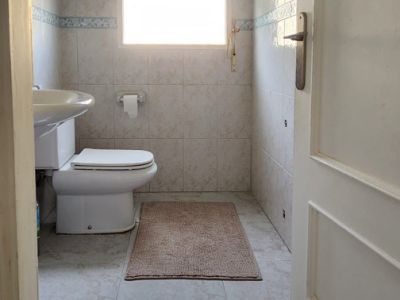 Baño funcional con inodoro, lavabo y ventana. Presenta azulejos de pared y suelo en tonos crema con un detalle decorativo azul, y una alfombrilla.