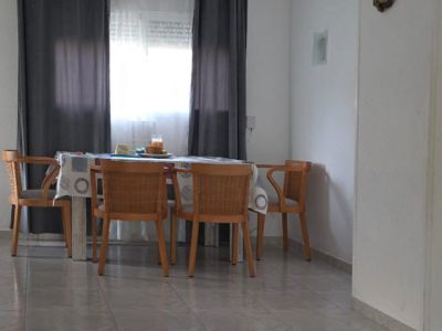 Comedor luminoso con mesa de madera y sillas de mimbre, junto a una ventana con cortinas grises. El suelo es de baldosas claras, complementado por una alfombra rayada en primer plano.