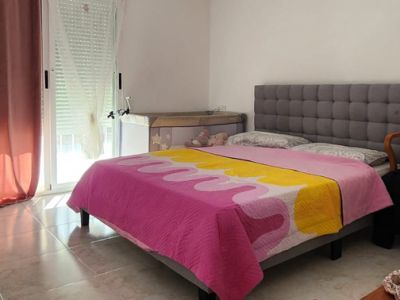 Dormitorio con cama tapizada en gris, suelo de baldosas claras, luz natural de ventana con persiana y una cuna. Equipado con aire acondicionado de pared.