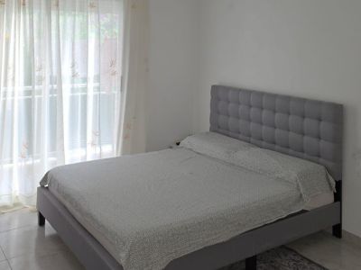 Dormitorio con cama tapizada gris y cabecero capitoné, con ropa de cama clara. Luz natural entra por el gran ventanal con visillos y cortinas granates, sobre suelo de baldosas.
