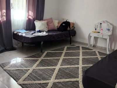 Dormitorio con suelo de baldosas claras, una alfombra geométrica gris y un diván. La ventana con cortinas oscuras permite la entrada de luz natural, junto a un tocador blanco.