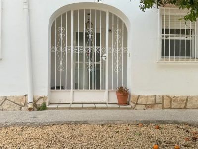 Entrada principal de la villa con un portón de metal arqueado y seguro. El jardín de grava frontal muestra frutos caídos y un árbol frutal que sombrea la fachada.