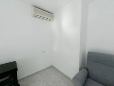 Esquina de una habitación con paredes blancas y suelo de baldosas claras. Dispone de aire acondicionado split y un enchufe en la pared, junto a un sillón gris y un mueble oscuro.