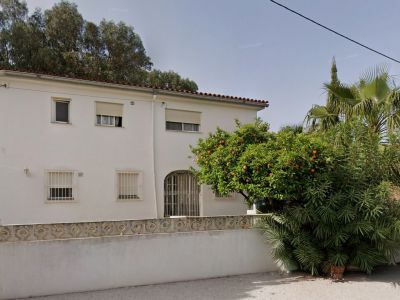 Fachada principal de la villa con paredes blancas, tejado de teja roja y ventanas con rejas. Destaca un naranjo cargado de fruta y exuberante vegetación en el jardín.