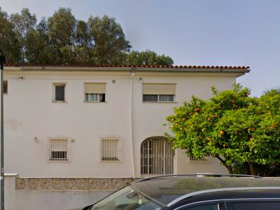La fachada blanca de la villa de dos plantas muestra un tejado tradicional, una entrada arqueada con reja ornamental y ventanas con rejas. Un naranjo maduro, cargado de frutos, se alza prominentemente.