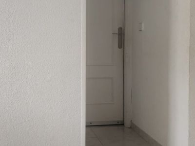 Pasillo interior con suelo de baldosas claras y paredes blancas texturizadas, mostrando una puerta panelada al fondo y un pequeño adorno en la pared.