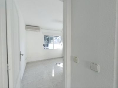 Vista de un dormitorio vacío con suelo de baldosas claras, paredes blancas y un aparato de aire acondicionado. La ventana con rejas de seguridad permite la entrada de luz natural.