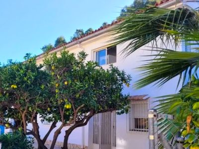 Vista exterior de la villa con fachada blanca, tejado de tejas y árboles frutales de cítricos con frutos amarillos. Se aprecian ventanas y una entrada.