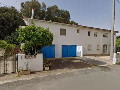 Vista frontal de la villa pareada con fachada blanca, dos puertas de garaje azules y un frondoso naranjo junto a la verja de hierro forjado que da acceso a la propiedad.